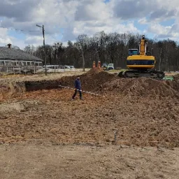 Wykop pod fundament domu z widoczną koparką i pracownikiem z poziomicą, na tle zabudowania gospodarczego i pochmurnego nieba.