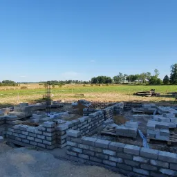 Fundamenty domu z bloczków betonowych na tle krajobrazu polnego w słoneczny dzień, widoczne zbrojenie i przygotowany wykop.