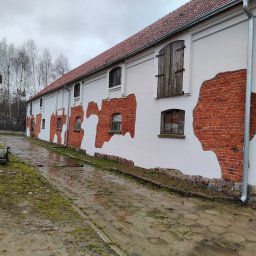 Firma Guzowski - Długi budynek z białymi ścianami i odsłoniętą czerwoną cegłą, widoczne drewniane okiennice i rynna, mokry brukowany podjazd, pochmurne niebo.