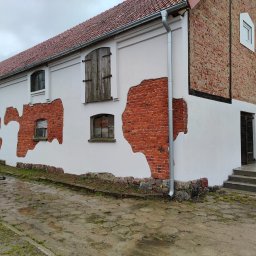 Firma Guzowski - Dwukondygnacyjny budynek z białymi ścianami i odsłoniętą czerwoną cegłą, z drewnianymi okiennicami, widoczny fragment dachu z czerwonej dachówki i mokry brukowany teren przed budynkiem.