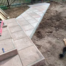 Układanie kostki brukowej na schodach i ścieżce ogrodowej, widoczny poziomica, gumowy młotek i nóż tapicerski.