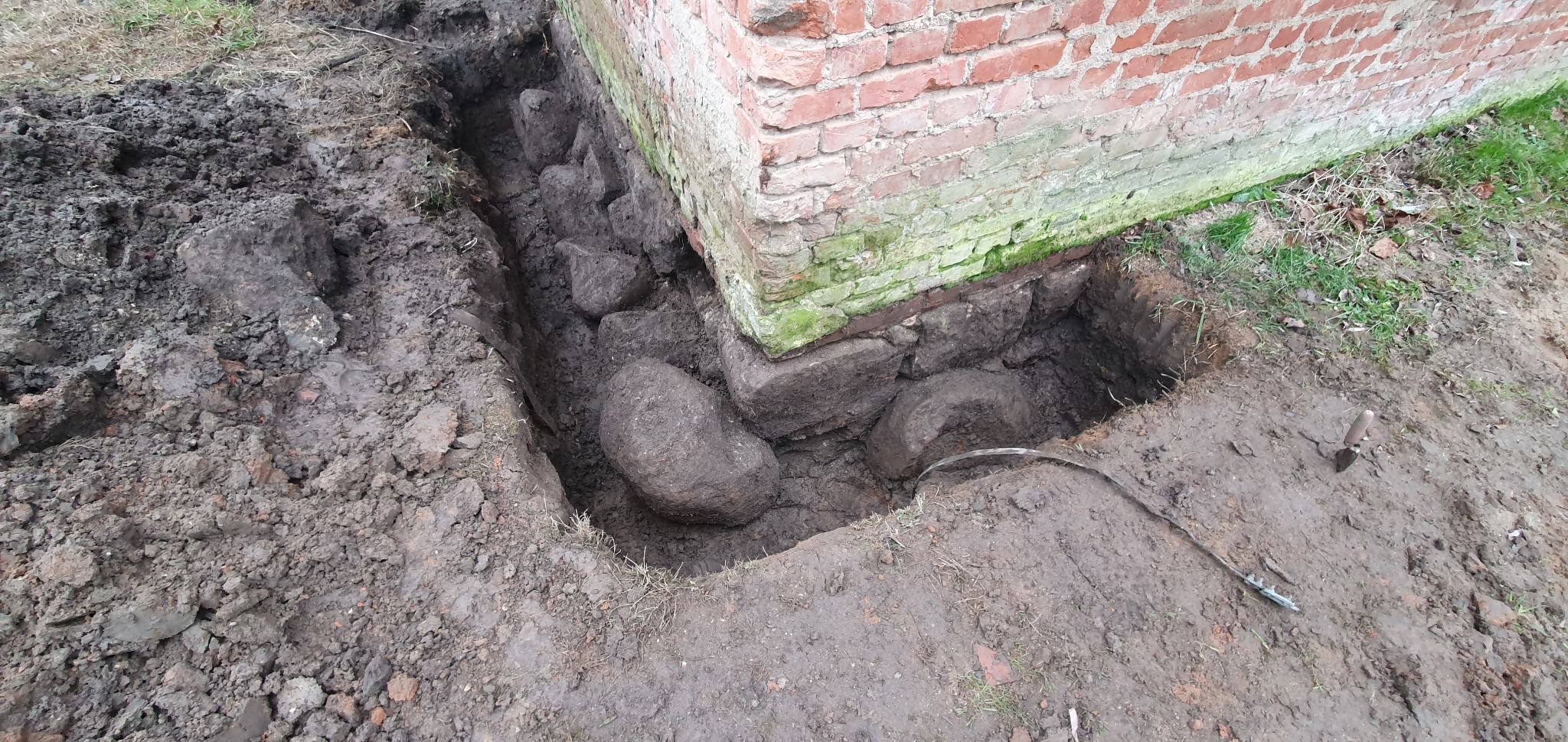 Odkopany fundament ceglanego budynku z widocznymi kamieniami, ziemią i narzędziem do kopania.