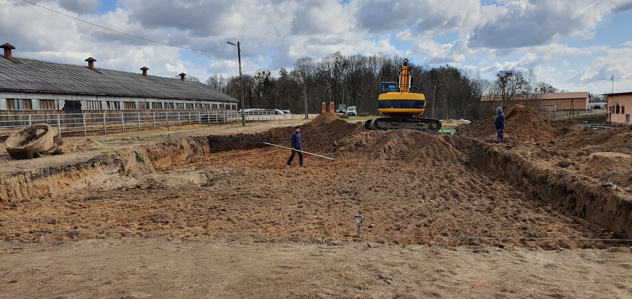 Wykop pod fundament domu z widoczną koparką i pracownikiem z poziomicą, na tle zabudowania gospodarczego i pochmurnego nieba.