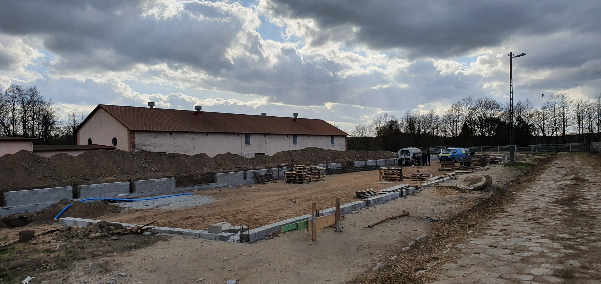 Fundamenty budynku gospodarczego w trakcie budowy, widoczne bloczki betonowe, wykop, hałdy ziemi, drewniane palety i zaplecze budowy z pojazdami na tle zachmurzonego nieba.