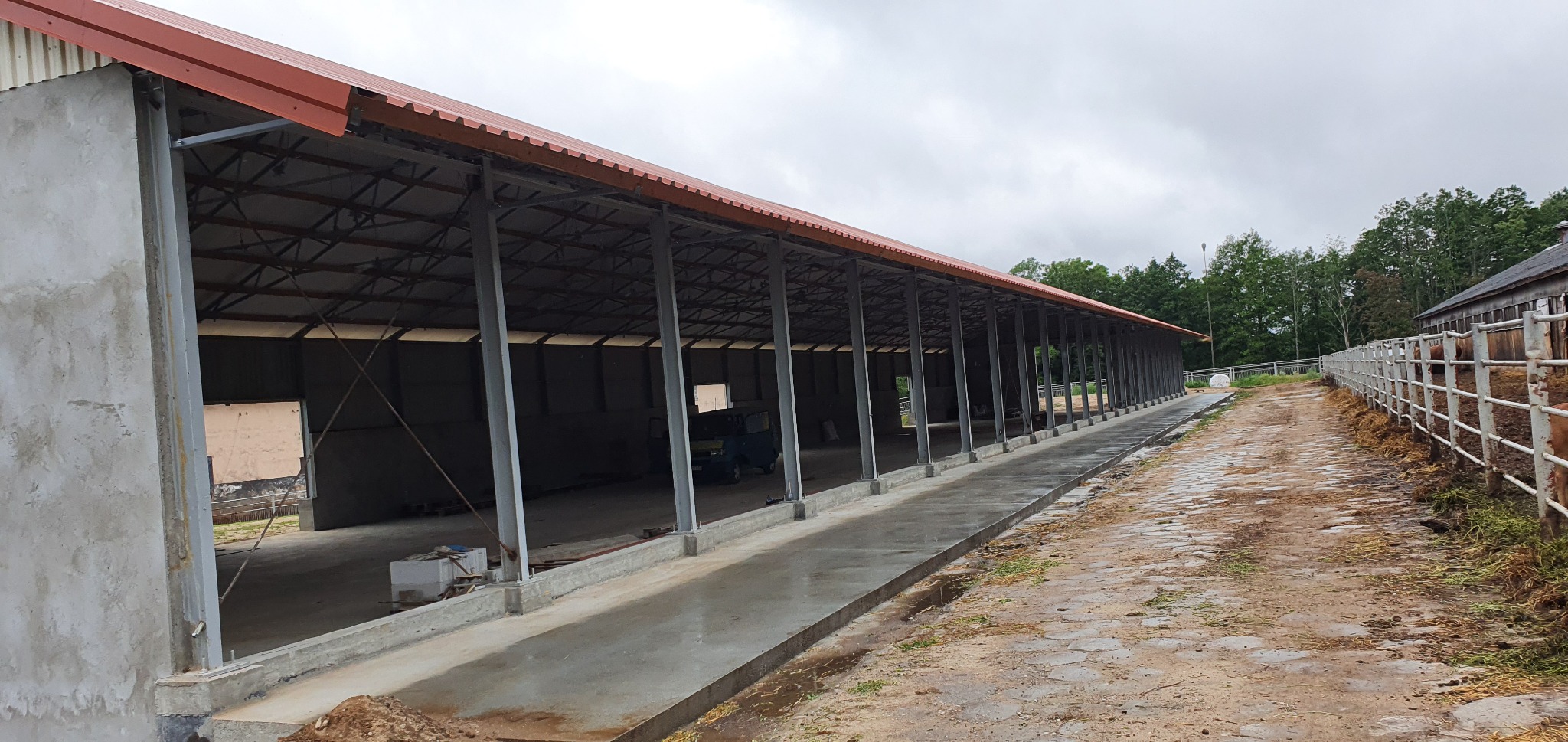 Konstrukcja hali z betonowymi ścianami, stalowymi słupami i czerwonym dachem z blachy trapezowej, widok z zewnątrz na tle pochmurnego nieba i drzew.