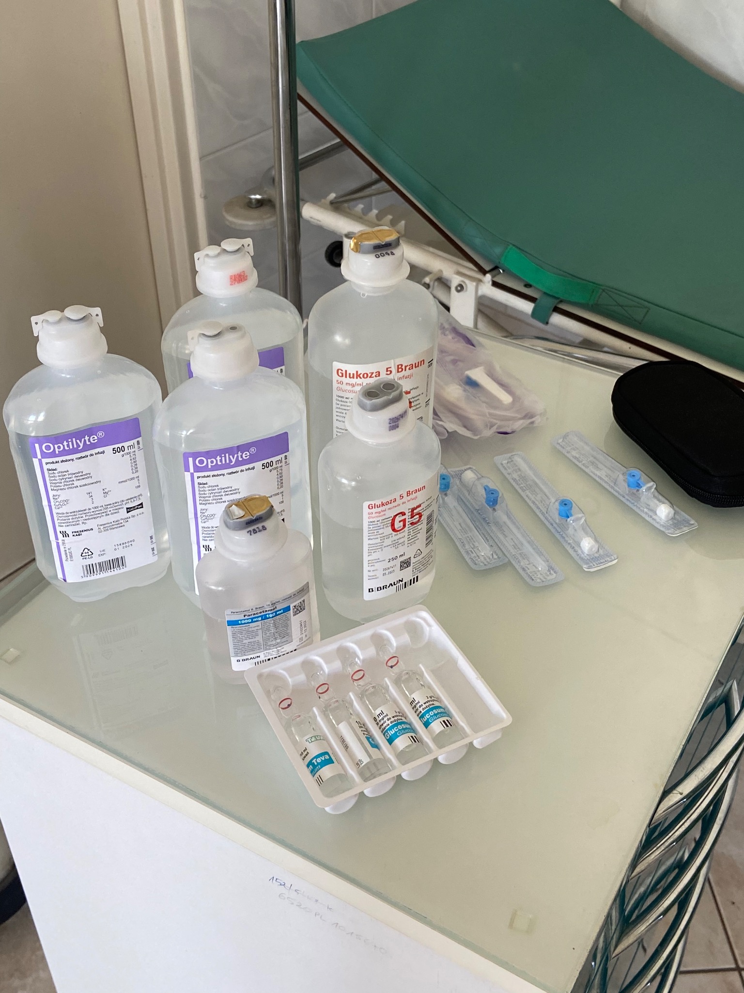 Butelki z roztworami do infuzji Optilyte i Glukoza 5 Braun oraz ampułki Teva i zestaw dożylny na stoliku medycznym z zielonym leżankiem w tle.