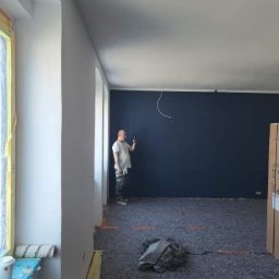 Piotr Makowski - Mężczyzna w t-shircie i spodniach roboczych stoi w pokoju z pomalowaną na granatowo ścianą, trzymając w ręku telefon, podłoga zabezpieczona folią i kartonem, widoczne ślady po taśmie malarskiej.