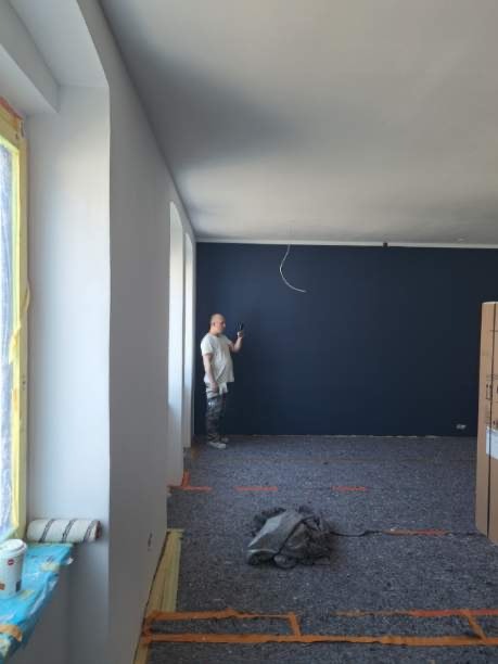 Mężczyzna w t-shircie i spodniach roboczych stoi w pokoju z pomalowaną na granatowo ścianą, trzymając w ręku telefon, podłoga zabezpieczona folią i kartonem, widoczne ślady po taśmie malarskiej.