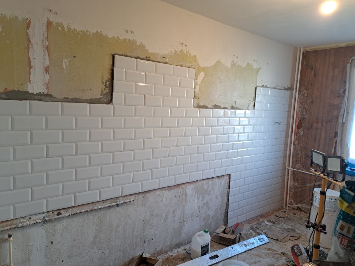 Układanie białych, prostokątnych płytek typu metro na ścianie, widoczne niedokończone fragmenty ściany z resztkami starej zaprawy, poziomica i lampa halogenowa na trójnogu w pomieszczeniu.