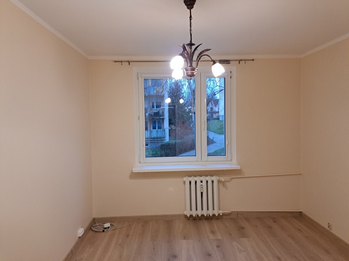 Pomalowane na beżowo puste wnętrze pokoju po remoncie z drewnopodobną podłogą, białym grzejnikiem pod oknem i ozdobnym żyrandolem.