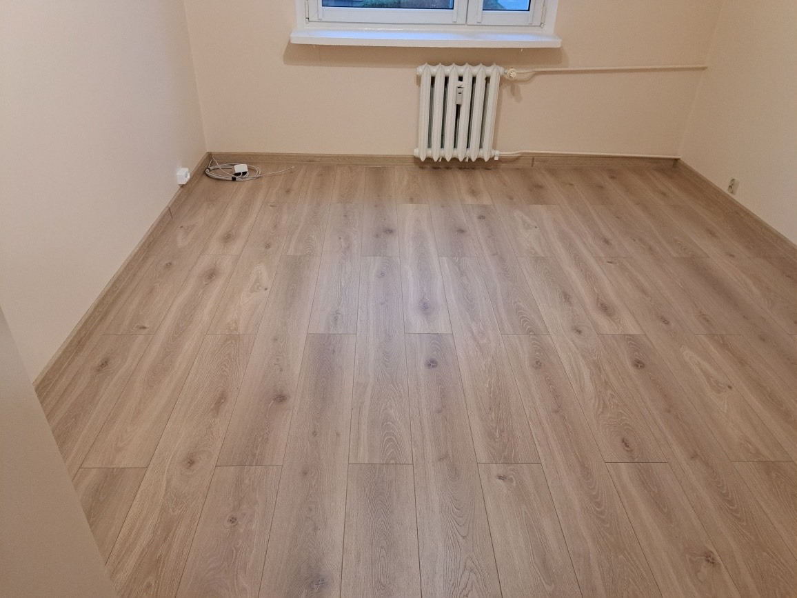 Wykończone wnętrze pokoju z panelami podłogowymi w odcieniach jasnego drewna, biały grzejnik pod oknem i beżowe ściany.