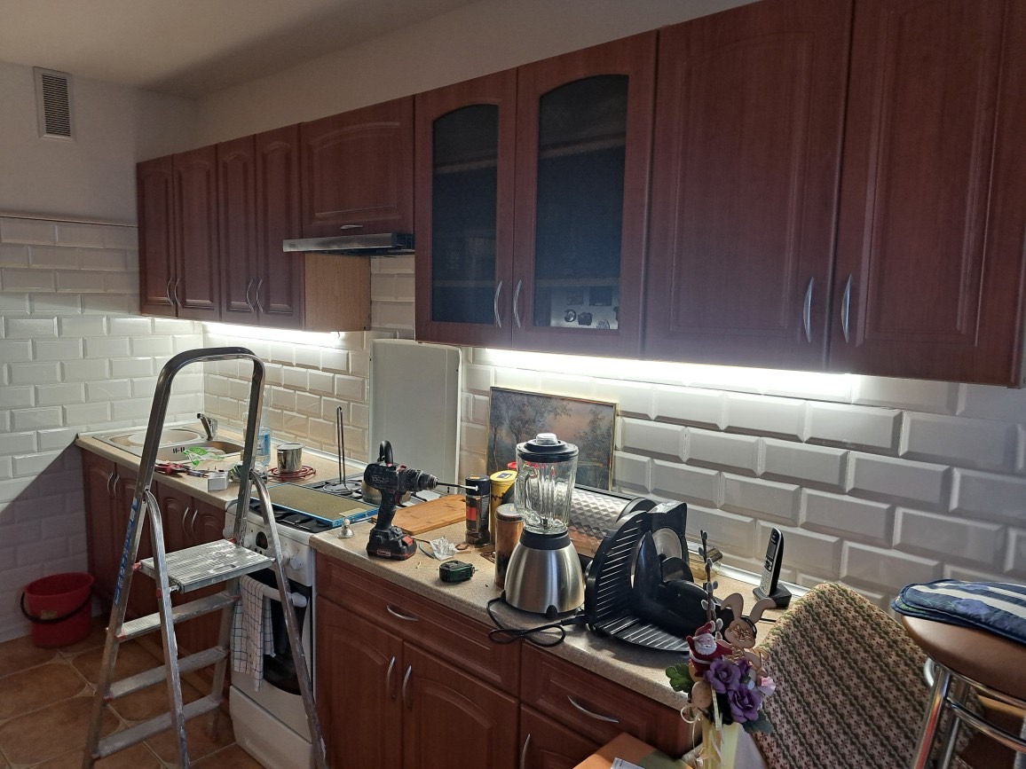 Kuchnia w trakcie prac: widoczne brązowe szafki, białe płytki typu metro, drabina aluminiowa, sprzęt AGD, wiertarka akumulatorowa na blacie, oraz oświetlenie LED pod szafkami wiszącymi.