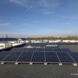 Instalacja fotowoltaiczna na płaskim dachu budynku z widocznym miastem w tle, wykonana w słoneczny dzień.