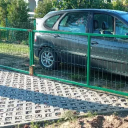 Zielona siatka ogrodzeniowa z bramą, widoczna na tle szarego samochodu i ażurowej kostki brukowej z kamieniami.