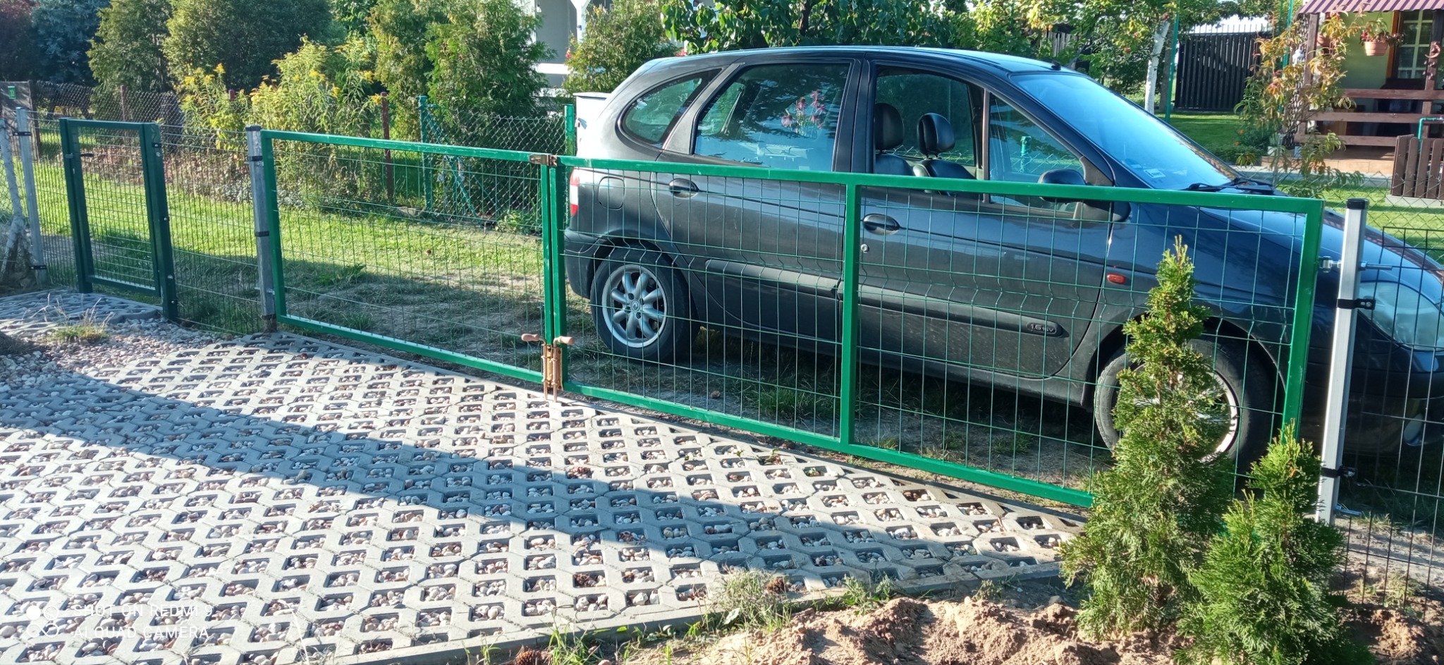 Zielona siatka ogrodzeniowa z bramą, widoczna na tle szarego samochodu i ażurowej kostki brukowej z kamieniami.