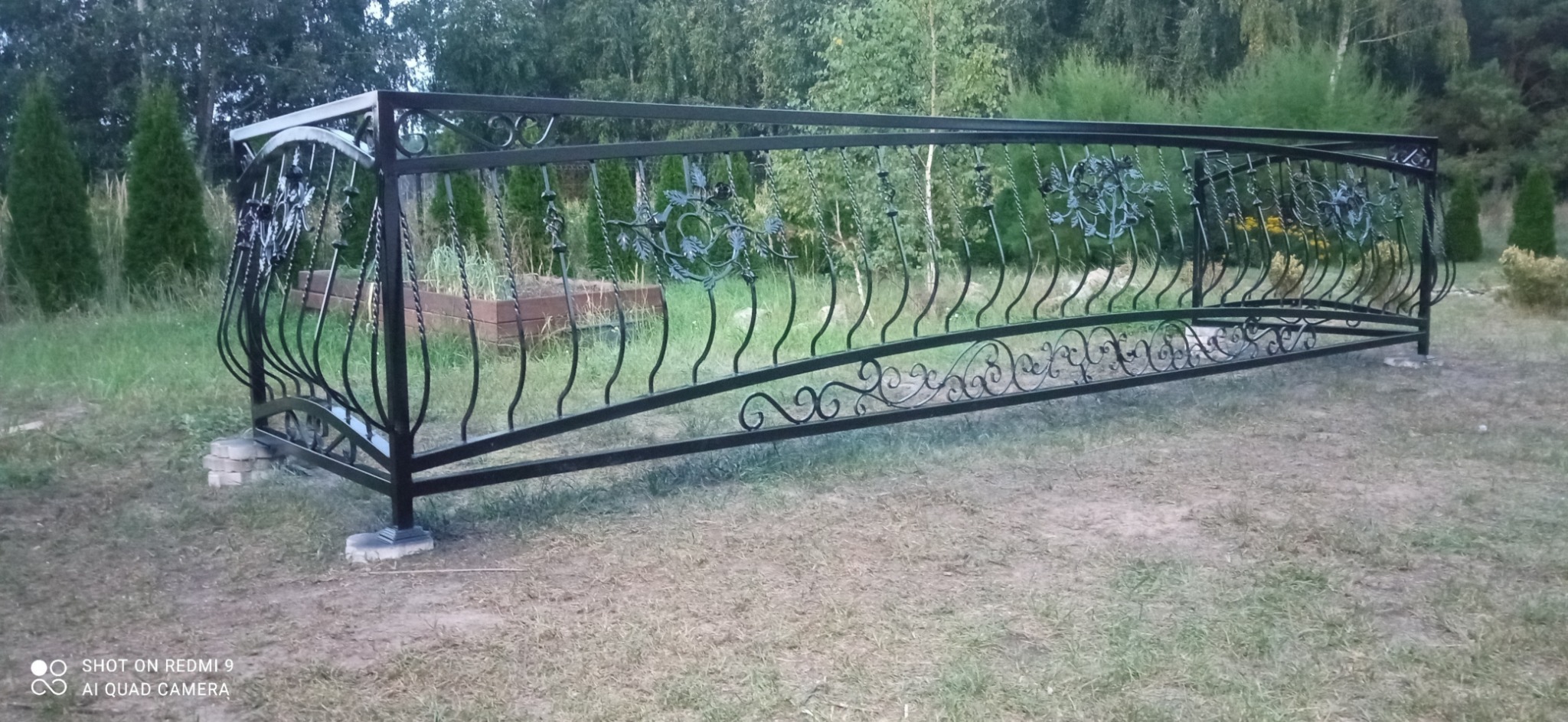 Czarna, metalowa balustrada z ozdobnymi motywami roślinnymi, zamontowana na niskiej podmurówce z cegieł na trawniku, z widocznymi drzewami i krzewami w tle.