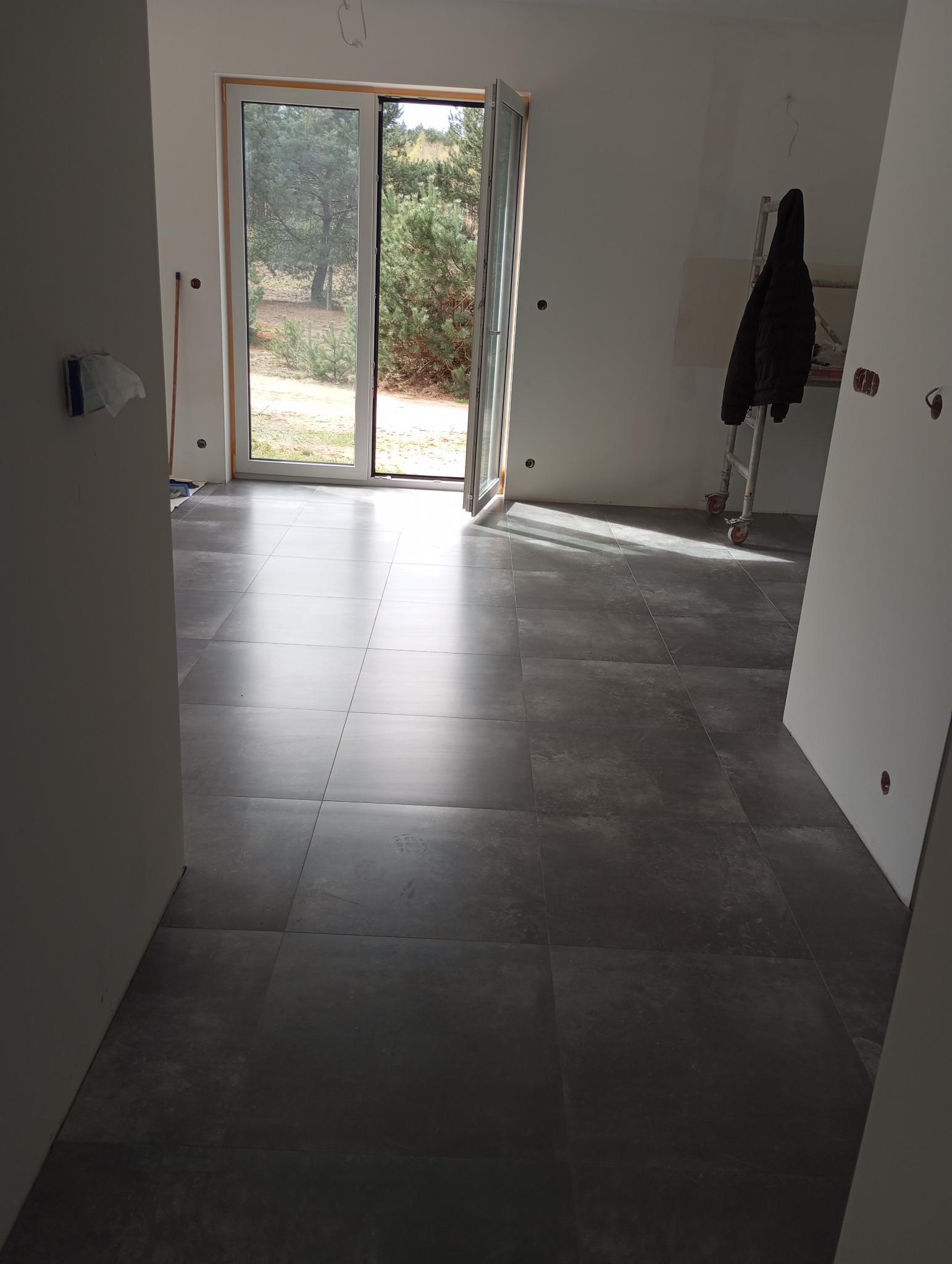 Wnętrze w trakcie wykańczania, z ułożoną, ciemnoszarą, kwadratową podłogą z płytek, widoczne otwarte drzwi balkonowe z widokiem na zewnątrz, drabina aluminiowa z wiszącą kurtką, ściany przygotowane...