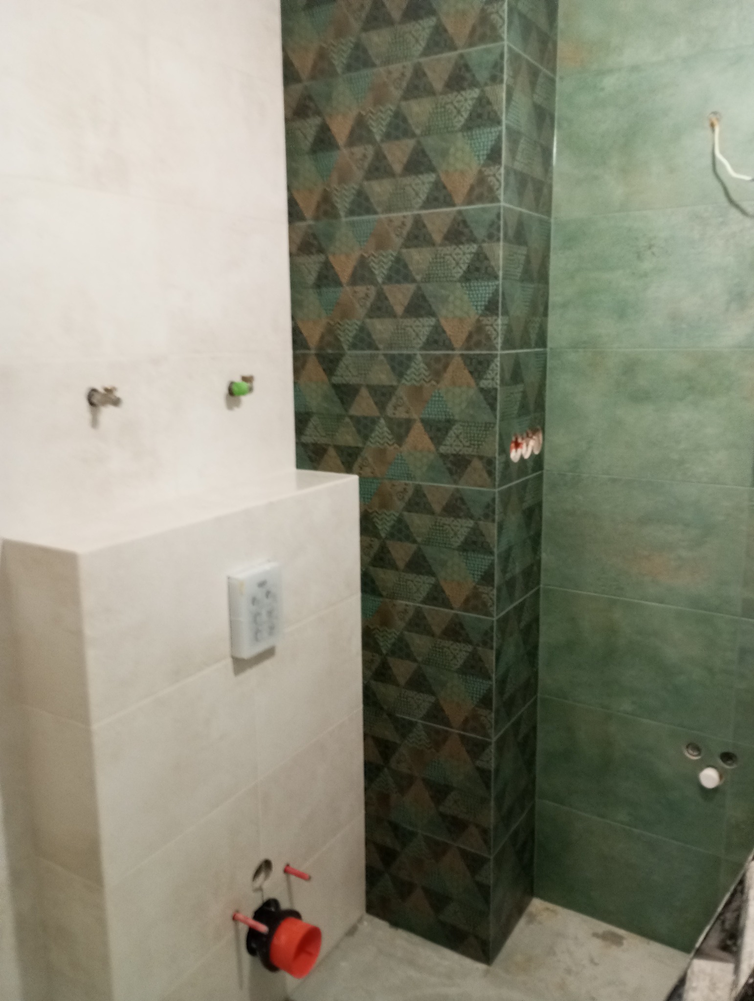 Wnętrze łazienki w trakcie remontu: widoczna zabudowa stelaża WC z przyciskiem spłukującym, fragment kolumny z płytek w geometryczny wzór, oraz zielone płytki na ścianie z wyprowadzonymi instalacjami.