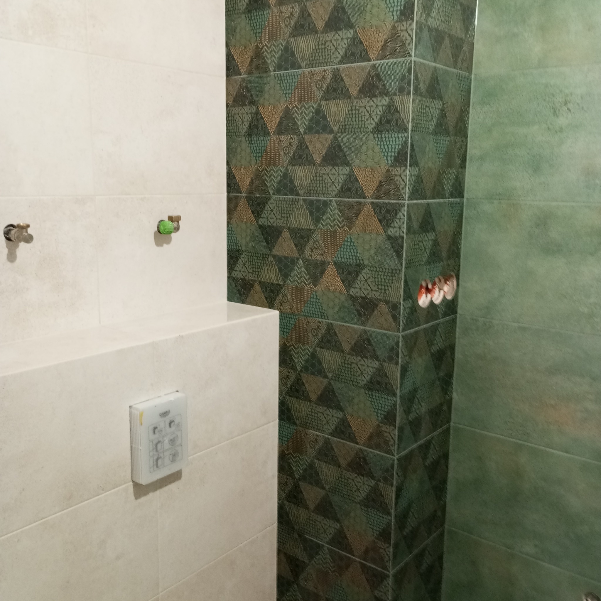 Fragment łazienki w trakcie wykańczania: beżowe i zielone płytki na ścianach, widoczne przyłącza wodne i przycisk spłuczki Grohe.