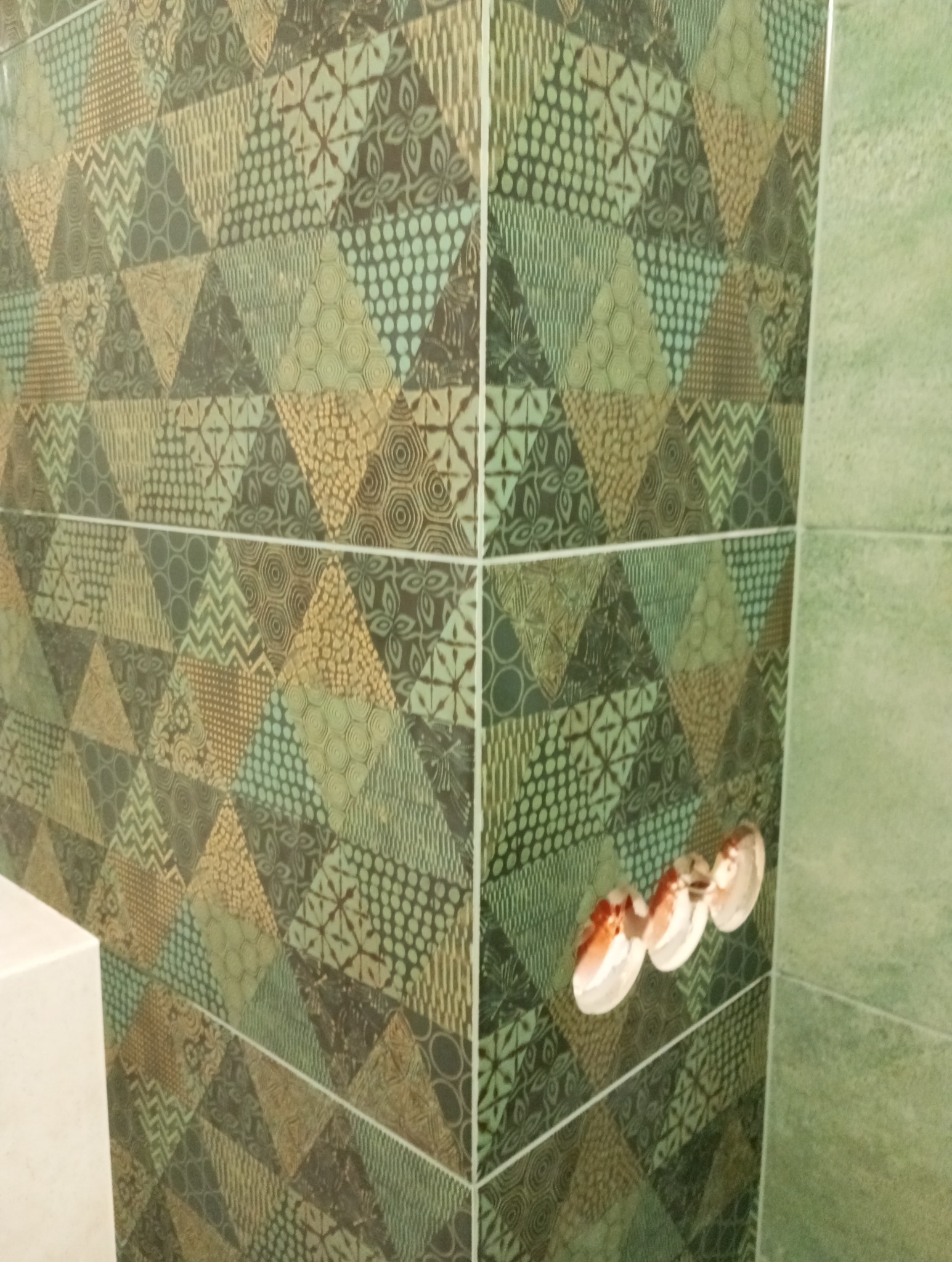 Ściana łazienki wyłożona zielono-brązowymi płytkami o geometrycznym wzorze, widoczne gniazdka elektryczne z białymi osłonami.