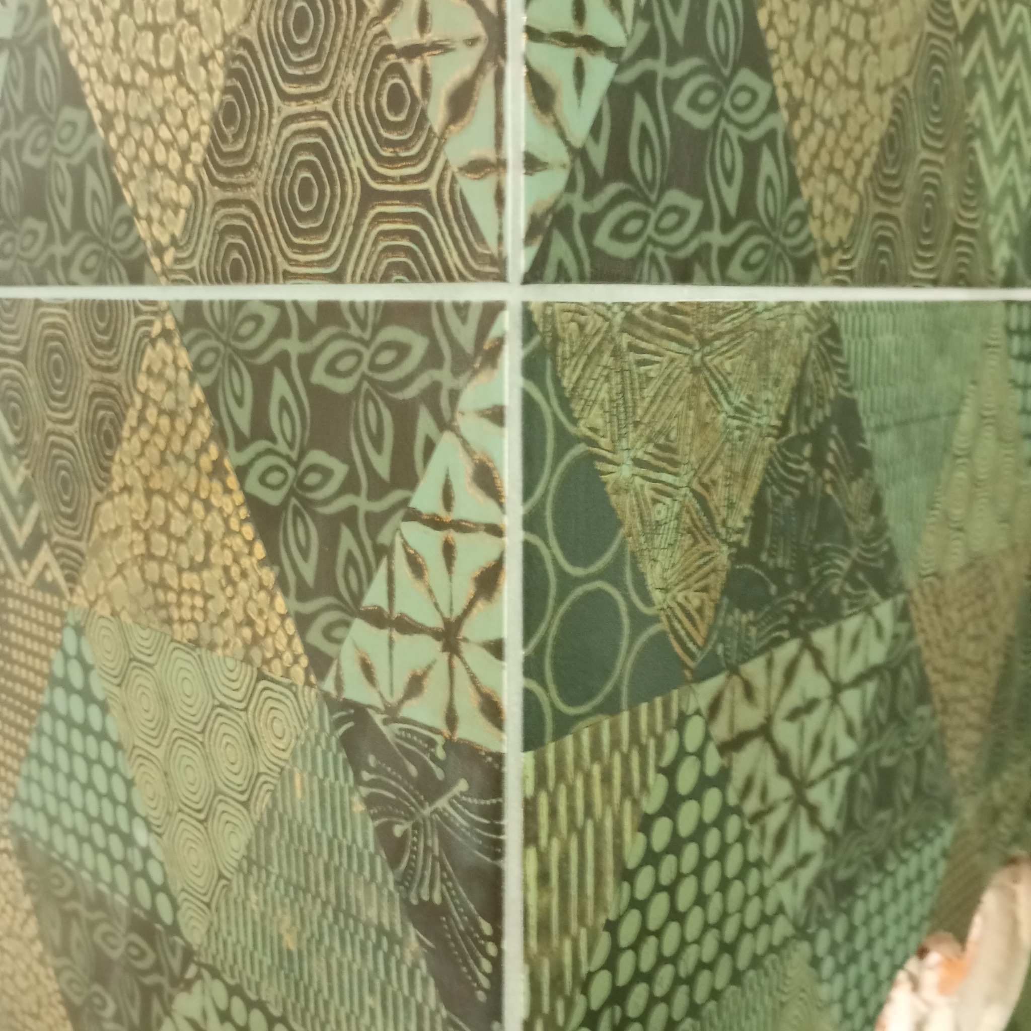 Fragment ściany wyłożonej dekoracyjnymi, zielono-beżowymi płytkami ceramicznymi o różnych wzorach geometrycznych i roślinnych, oddzielonych białymi fugami.