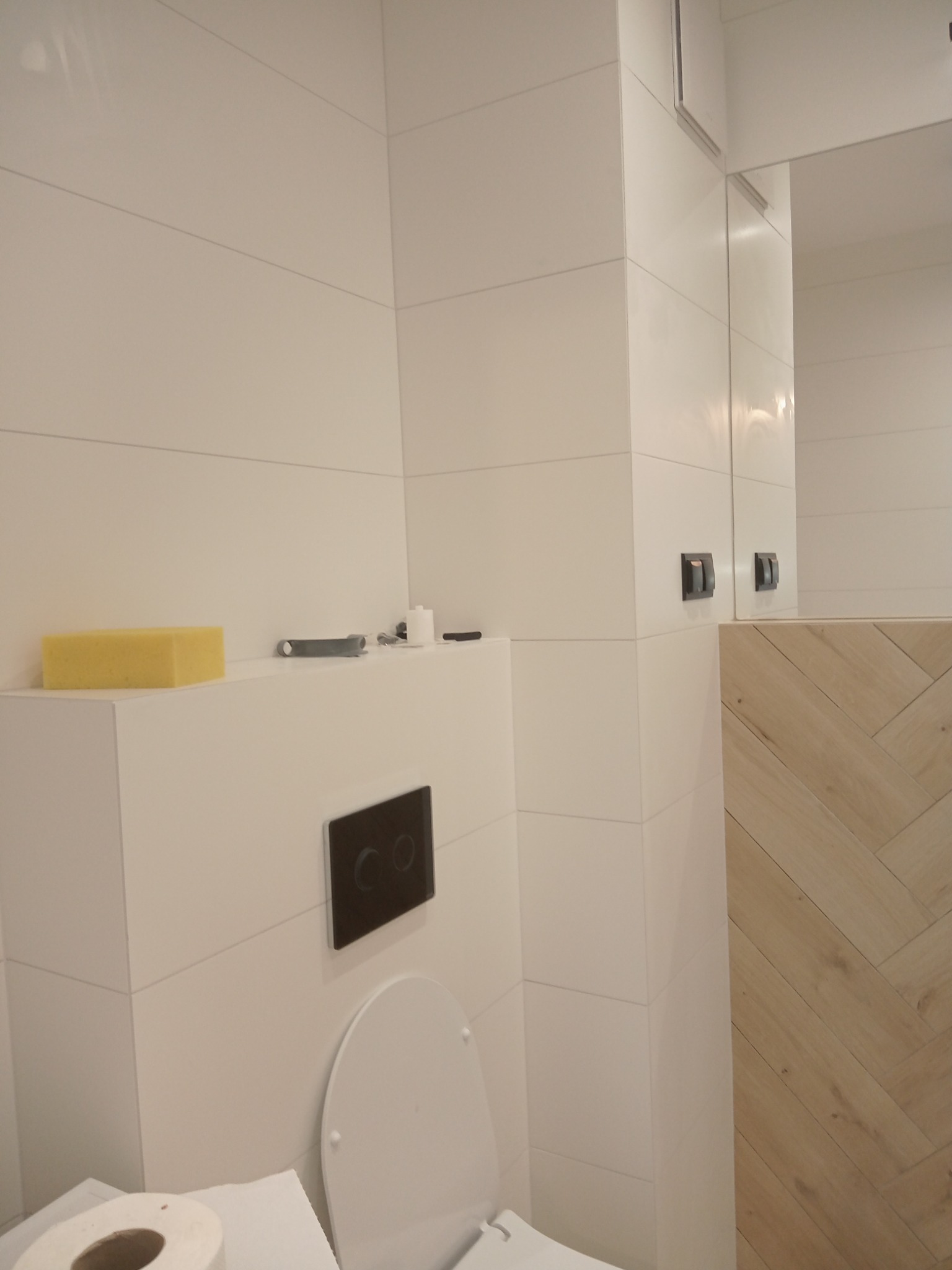 Nowoczesna łazienka z białymi, poziomymi płytkami, drewnianą ścianą w jodełkę, podwieszaną toaletą z czarnym przyciskiem i żółtą gąbką na obudowie stelaża.