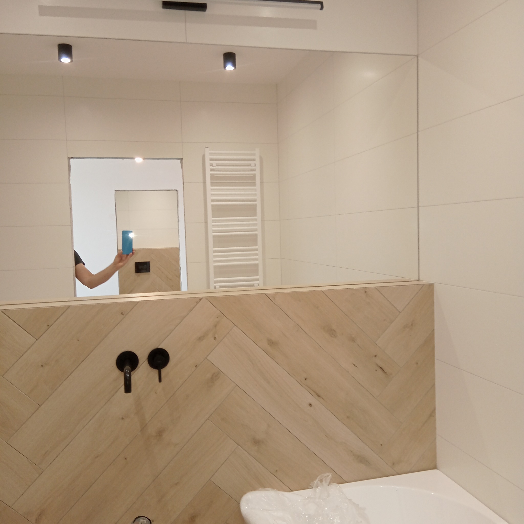 Nowoczesna łazienka z białymi płytkami, drewnopodobną zabudową w jodełkę, czarną armaturą i dużym lustrem, w którym odbija się osoba robiąca zdjęcie telefonem.