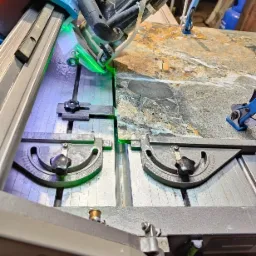 Cięcie płytki gresowej na mokro przy użyciu przecinarki elektrycznej z laserowym wskaźnikiem, widoczne zaciski mocujące i fragment blatu z ułożonymi płytkami.