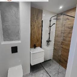 Nowoczesna łazienka z kabiną prysznicową typu walk-in, umywalką z szafką i toaletą w kolorze białym, szare płytki podłogowe z marmurkowym wzorem, ściany wykończone płytkami w odcieniach bieli...