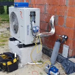 Biała jednostka zewnętrzna z logo Rotenso na betonowym fundamencie, podłączona do instalacji chłodniczej; obok narzędzia i butla z gazem, ściana z czerwonej cegły w tle.