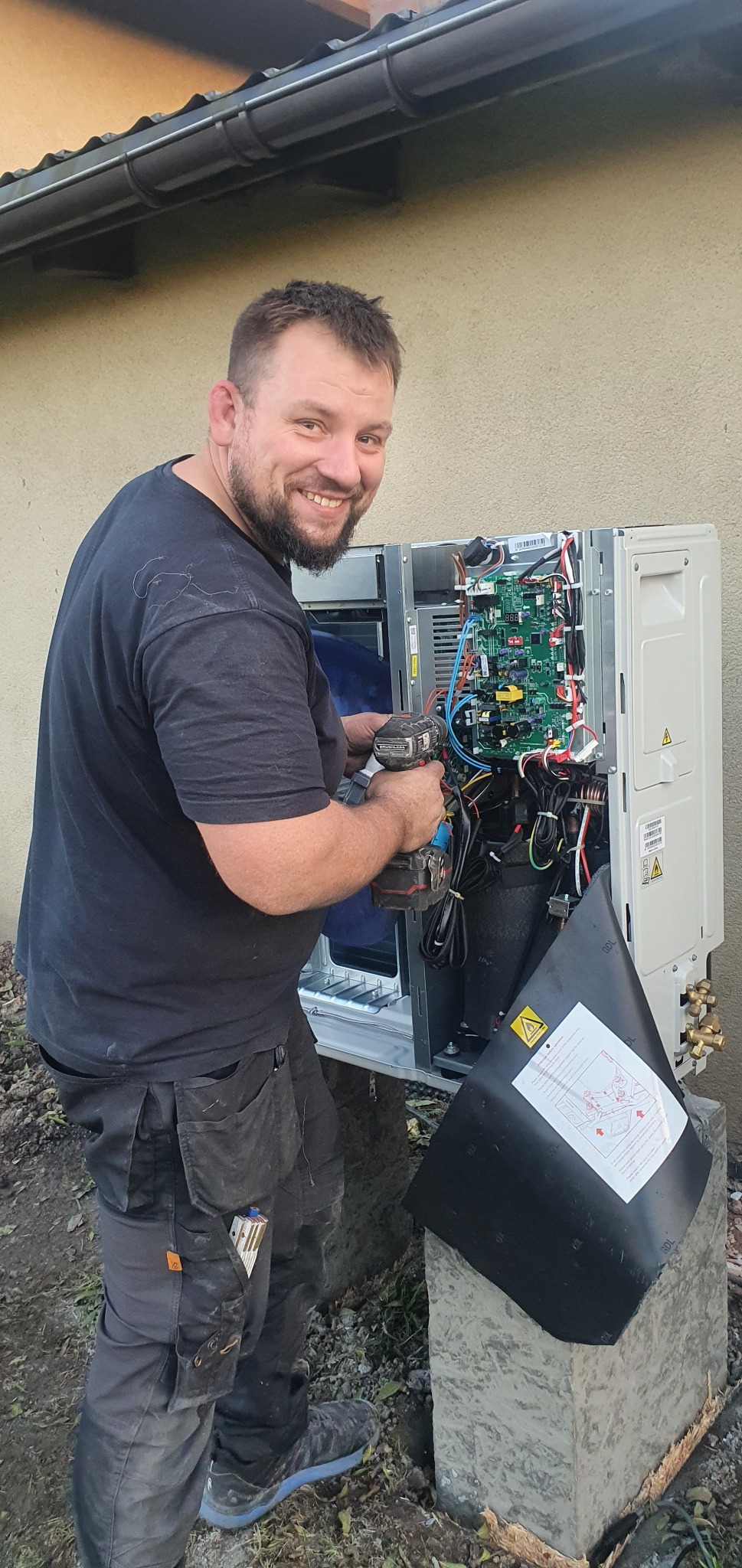 Uśmiechnięty instalator w czarnym ubraniu z wkrętarką w dłoni podczas montażu otwartej pompy ciepła na zewnątrz budynku, widoczna płyta sterująca i okablowanie.