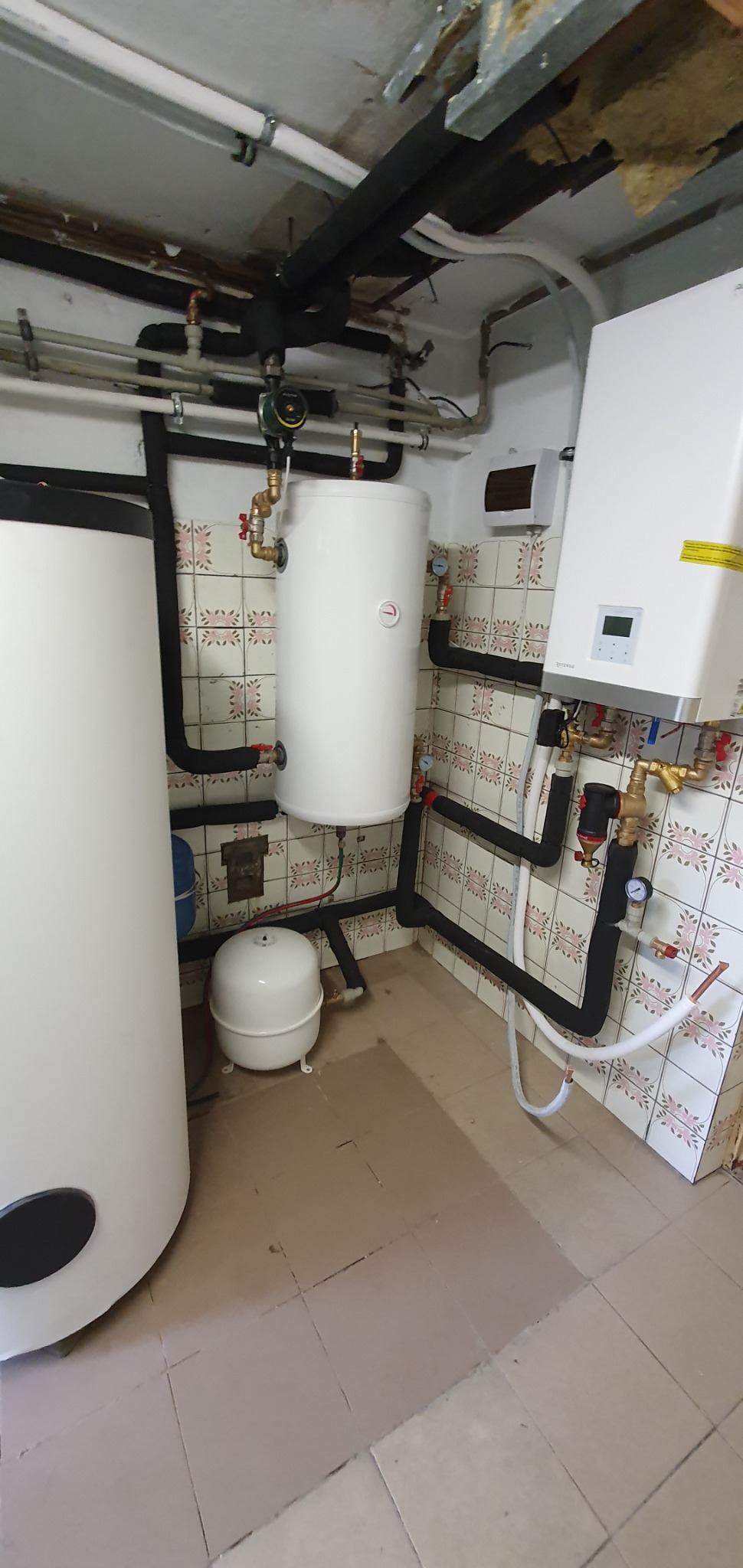 Kotłownia z dwoma białymi zbiornikami, małym zbiornikiem wyrównawczym, rurami w izolacji termicznej i płytkami ceramicznymi na ścianach.