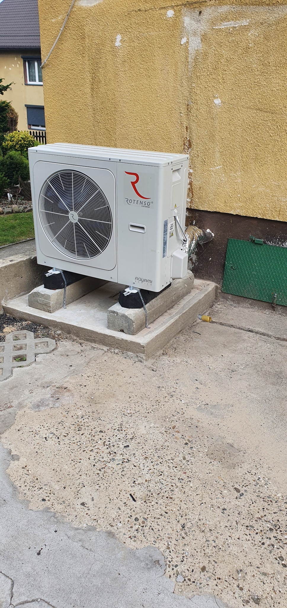 Biała pompa ciepła Rotenso Aquami, zamontowana na betonowym fundamencie z podkładkami antywibracyjnymi, z widoczną aluminiową rurą wentylacyjną przy żółtej, teksturowanej ścianie budynku.