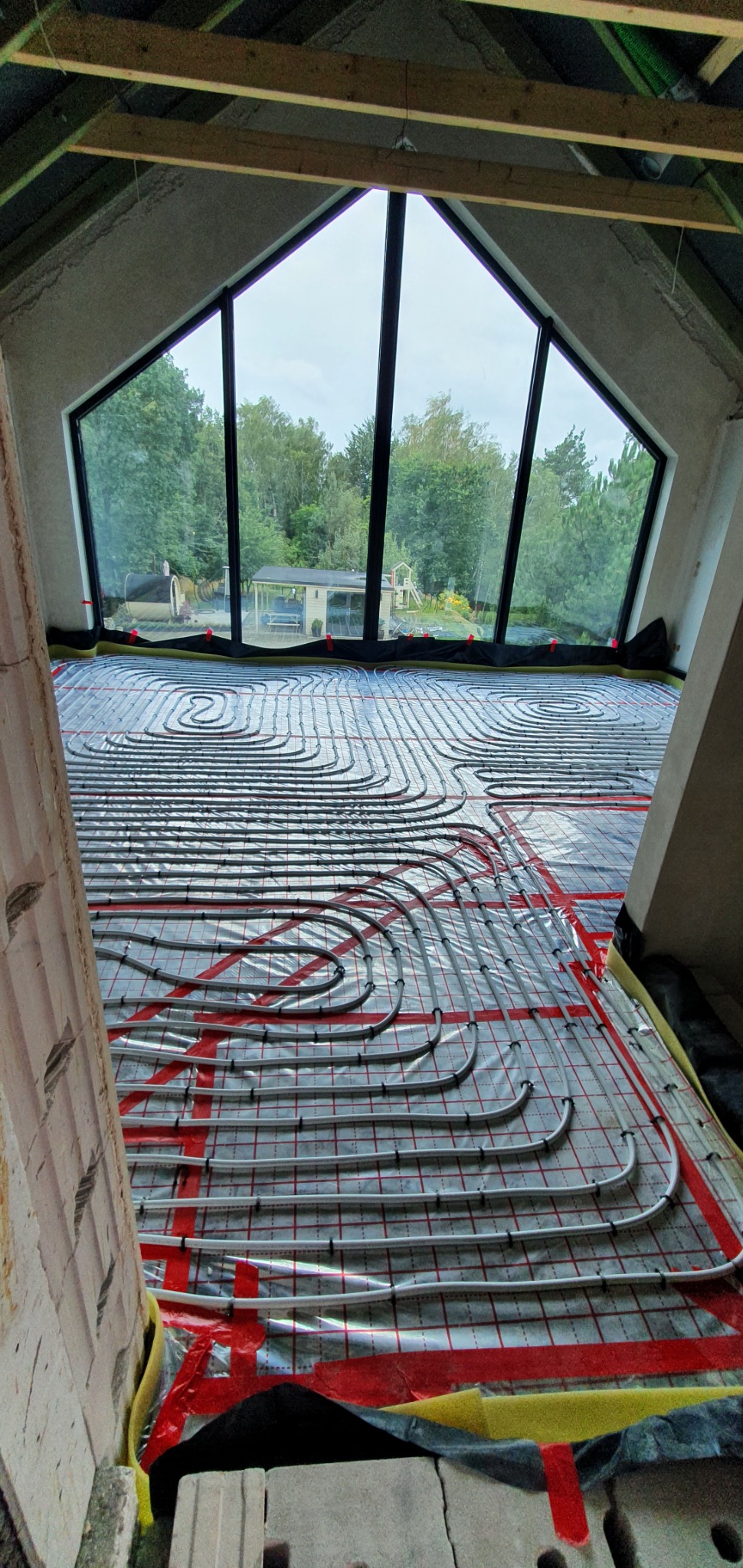 Układanie rur ogrzewania podłogowego w nowym domu z dużym trójkątnym oknem, widok na ogród z domkiem i placem zabaw.