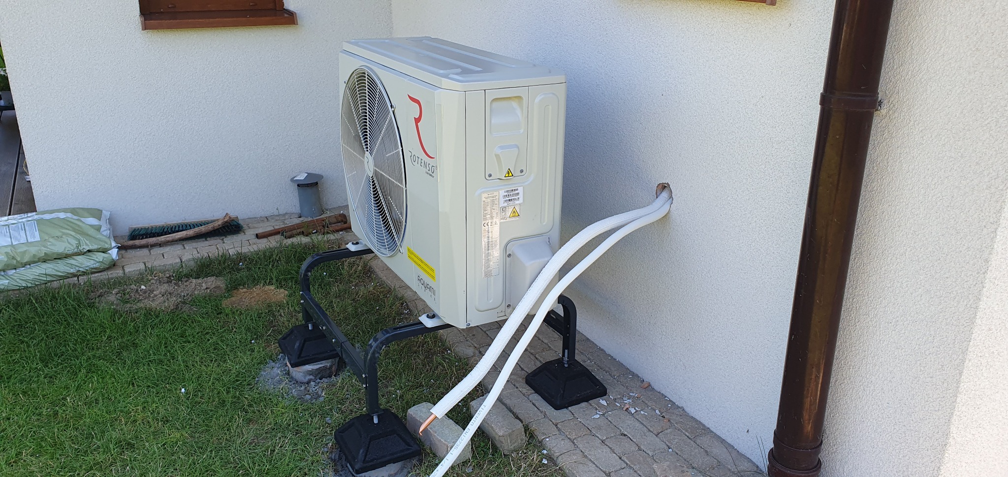 Zewnętrzna jednostka pompy ciepła Rotenso Aquami, zamontowana na czarnej, metalowej podstawie na trawniku przy ścianie domu z widocznymi rurami chłodniczymi w białej izolacji.
