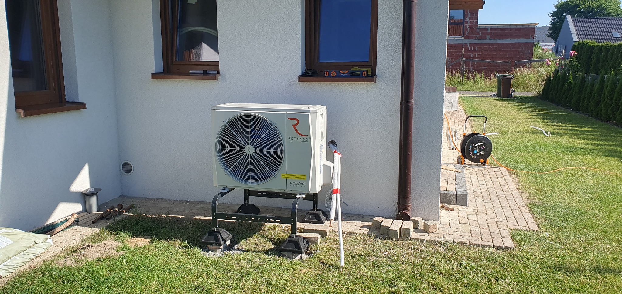 Biała jednostka zewnętrzna z logo Rotenso zamontowana na czarnym stelażu przy ścianie budynku z brązowymi oknami, widoczny fragment trawnika i kostki brukowej.