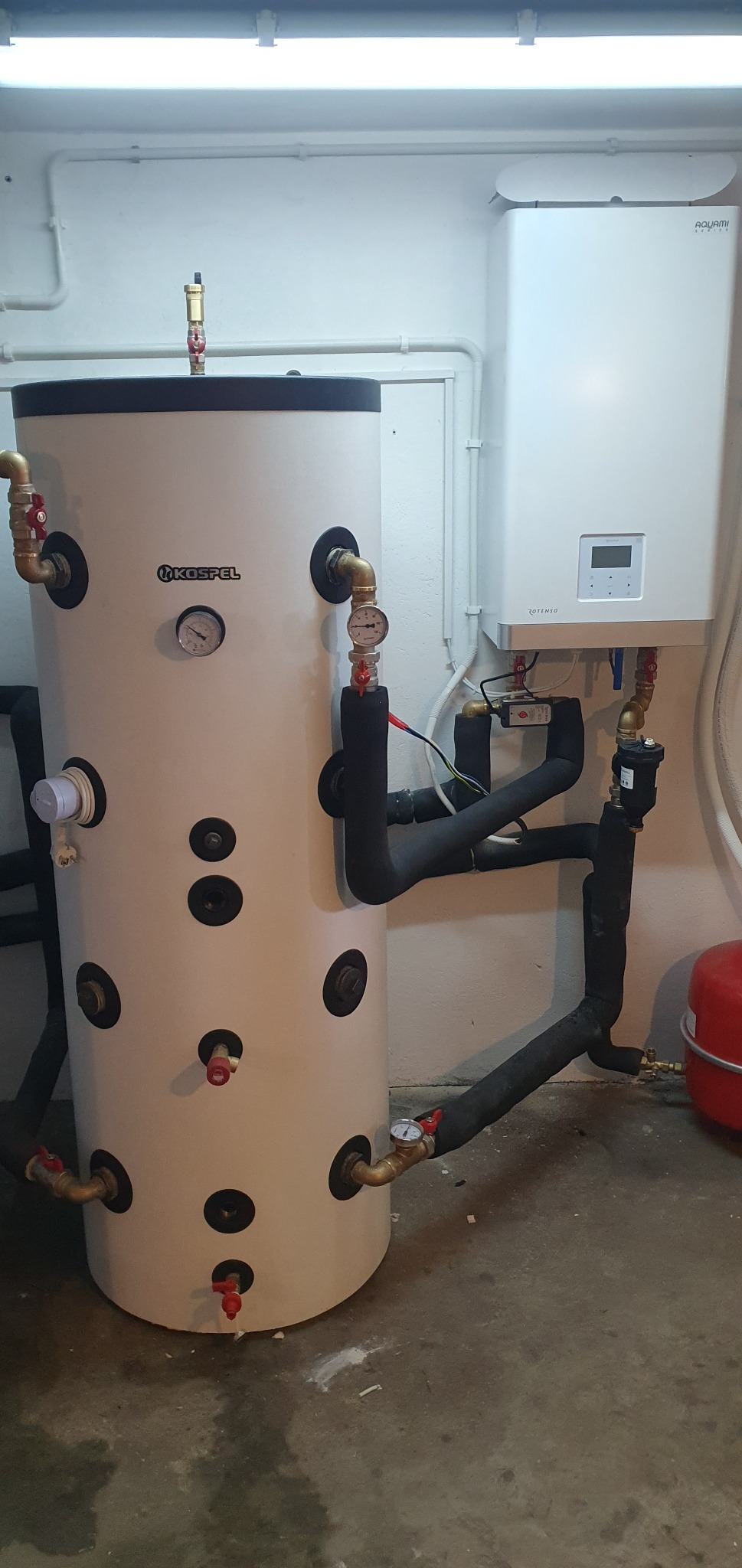 Biały zasobnik ciepłej wody użytkowej Kospel z widocznymi przyłączami i manometrami, obok wiszący piec gazowy Aquami Rotensa z wyświetlaczem, rury w izolacji termicznej, instalacja w kotłowni.