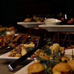 Maya Heller - Elegancko zastawiony stół z przekąskami: roladki, mini burgery z dekoracją z kiełków i listków, sosy w białych miseczkach, szczypce do nakładania, ciemne tło.