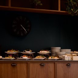 Maya Heller - Stół bufetowy z przekąskami na białych talerzach z metalowymi podstawkami, stosy białych talerzy, czarny zegar na ciemnozielonej ścianie, drewniane szafki i rośliny w tle.