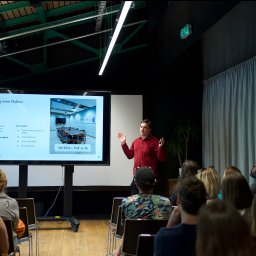 Maya Heller - Prelegent w czerwonej koszuli prezentuje ofertę sali konferencyjnej Meeting room Skyliner na ekranie, siedząca publiczność słucha