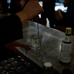 Maya Heller - Barman miesza drinka w szklance z lodem na marmurowym blacie baru, obok puszki napojów i butelka bitterów.