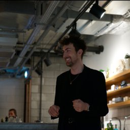 Maya Heller - Uśmiechnięty mężczyzna w czarnej marynarce, z krótkimi kręconymi włosami i złotym łańcuszkiem, w nowoczesnym wnętrzu z widocznymi instalacjami sufitowymi i półkami z dekoracjami.