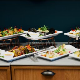 Maya Heller - Catering eventowy: różnorodne przekąski serwowane na białych tacach na metalowych stojakach, w tym mini burgery z dekoracyjnymi szpilkami, szaszłyki i czarne danie z limonkami, szczypce do nakładania.
