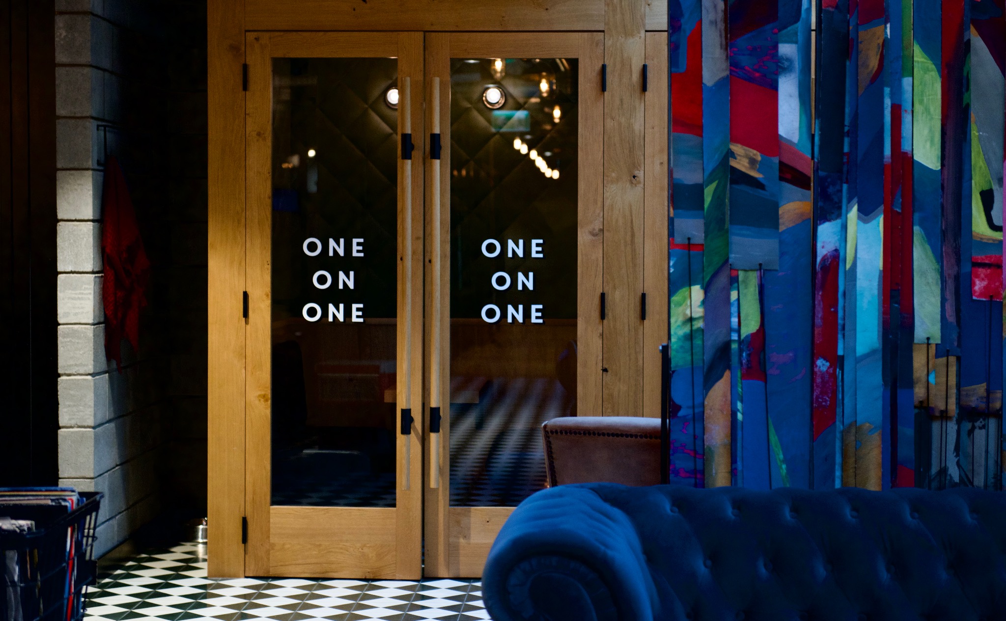 Wejście do lokalu z drewnianymi drzwiami z napisem 'ONE ON ONE', czarno-biała podłoga w szachownicę, fragment niebieskiej sofy i kolorowa ściana z drewnianych listew.