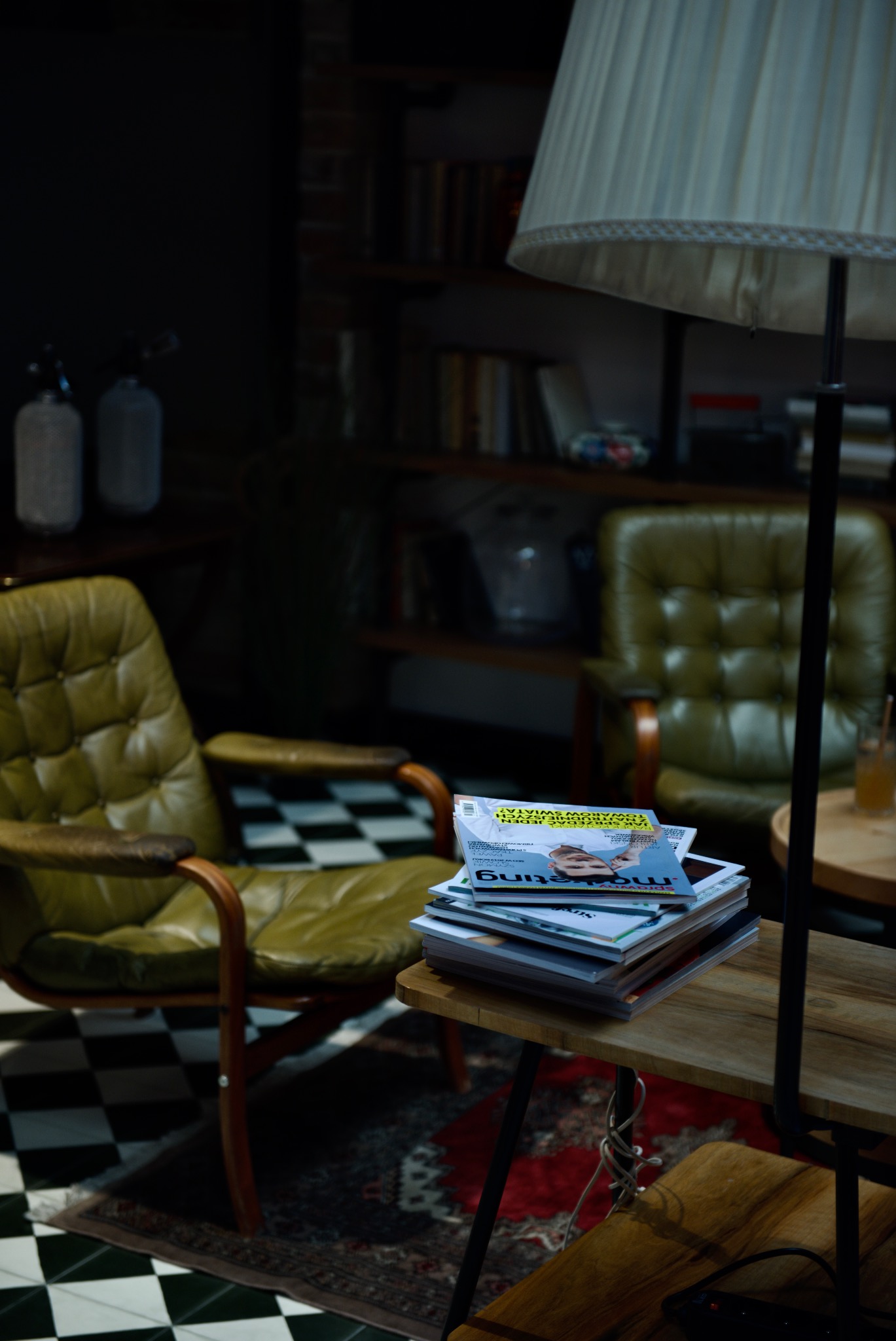 Stos magazynów na drewnianym stoliku obok zielonego skórzanego fotela w pomieszczeniu z szachownicową podłogą i regałem z książkami w tle.