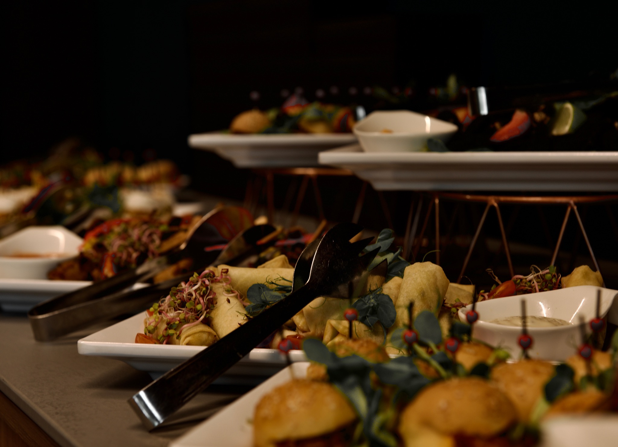 Elegancko zastawiony stół z przekąskami: roladki, mini burgery z dekoracją z kiełków i listków, sosy w białych miseczkach, szczypce do nakładania, ciemne tło.