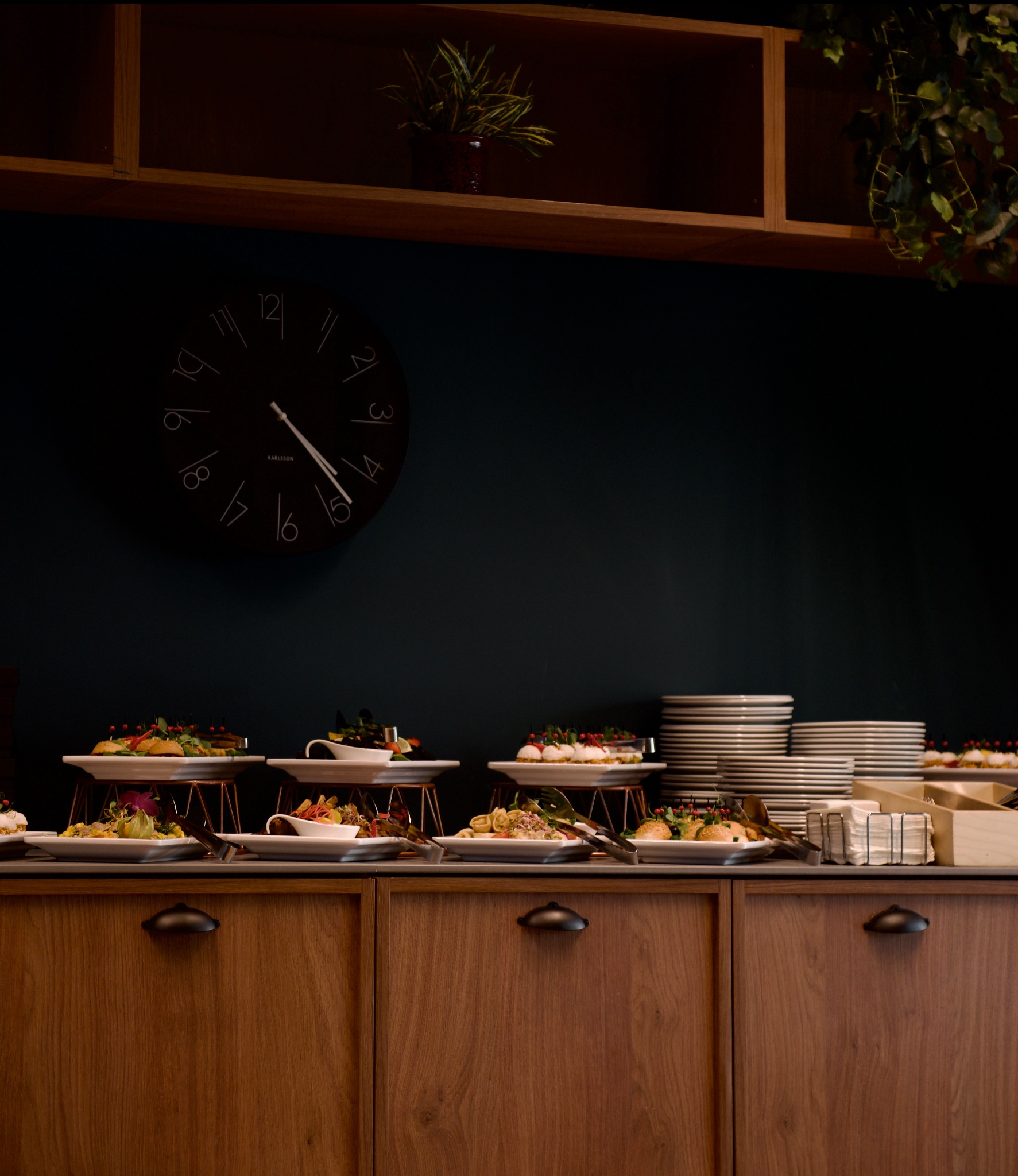 Stół bufetowy z przekąskami na białych talerzach z metalowymi podstawkami, stosy białych talerzy, czarny zegar na ciemnozielonej ścianie, drewniane szafki i rośliny w tle.