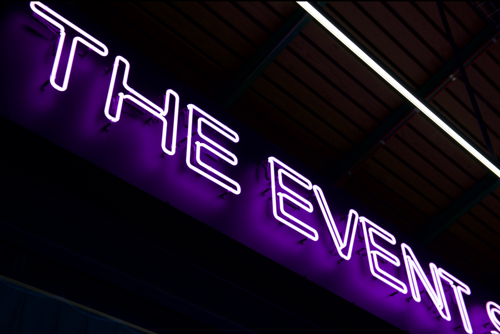 Neonowy napis 'THE EVENT' w fioletowym kolorze, widoczny pod kątem, na tle ciemnego sufitu z drewnianymi elementami i oświetleniem liniowym, Warszawa.