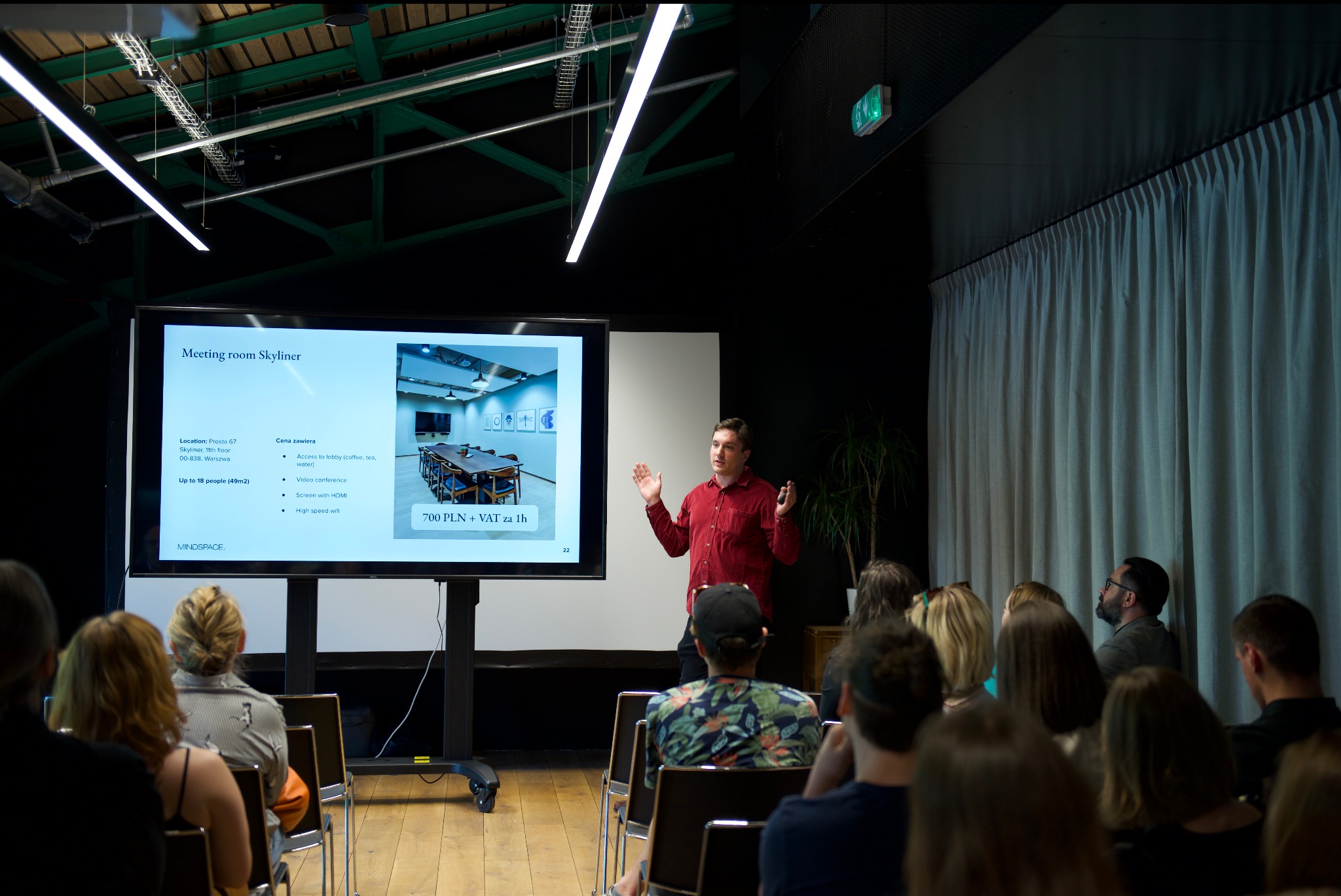 Prelegent w czerwonej koszuli prezentuje ofertę sali konferencyjnej Meeting room Skyliner na ekranie, siedząca publiczność słucha