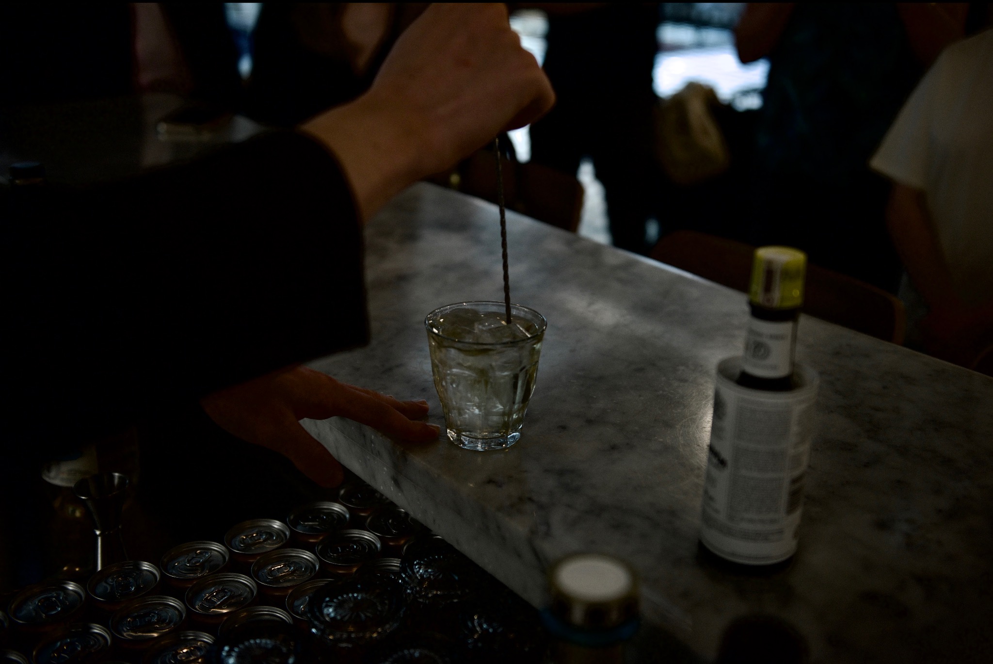 Barman miesza drinka w szklance z lodem na marmurowym blacie baru, obok puszki napojów i butelka bitterów.